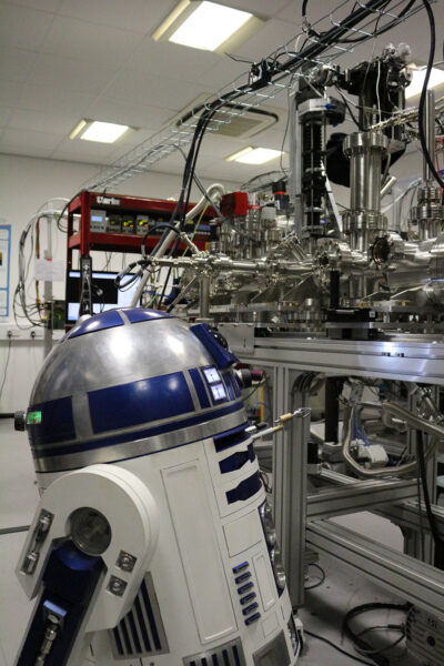 A real-size R2-D2 replica stands close to an instrument with a vacuum chamber and metallic components mounted on top of an optic table.