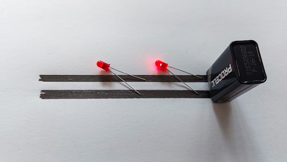 9 V battery standing on its contacts on two black lines drawn on a sheet of paper. Two red LEDs lying next to it, touching the lines with their contacts. One LED emitting red light.