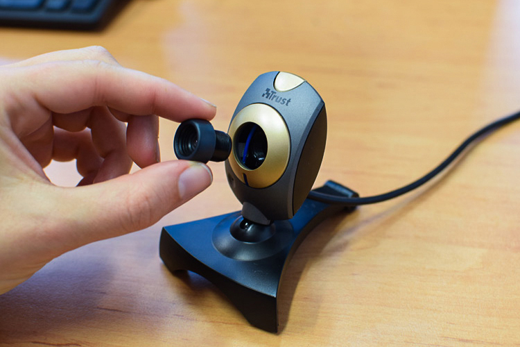 Hand pulling out the lens from a webcam that is standing on a wooden desk.