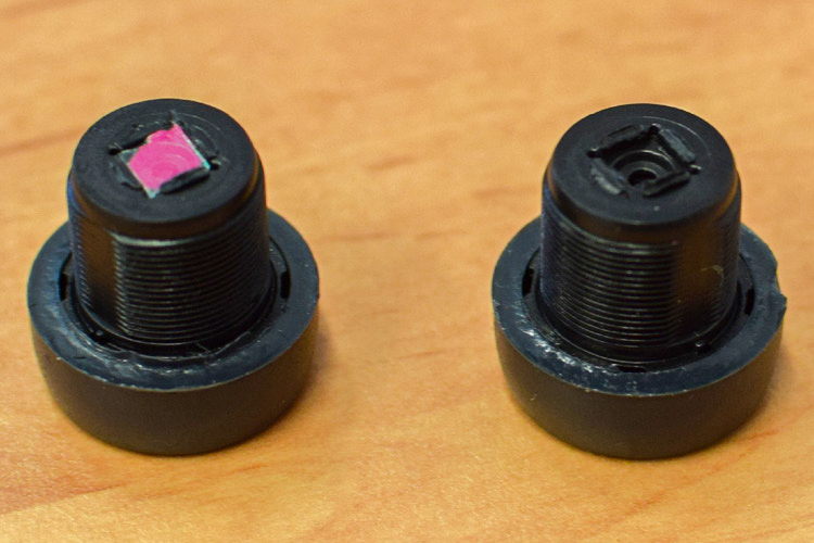 Two webcam lenses standing on a wooden desk. From one of them, a small pink plastic piece has been removed.