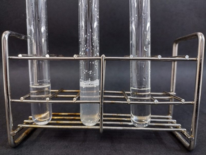 Porta provette con tre provette contenenti liquidi incolore. La provetta centrale contiene dei cristalli bianchi sulla superficie del liquido e lungo le pareti interne.