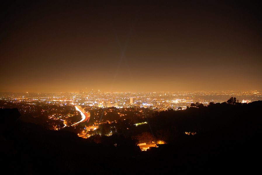 Panorama of a big city at night seen from a raised position in the distance. Illuminated buildings, streets, and a ‘light dome’ lightening up the sky above the city can be seen.