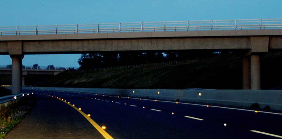 Concrete bridge spanning a road at night, with cat’s eyes on the road and reflectors on barriers along its sides.
