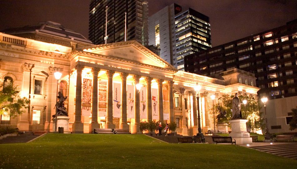 Fachada iluminada que representa un edificio con columnas. Hay un prado enfrente y edificios altos funcionales en el fondo. 