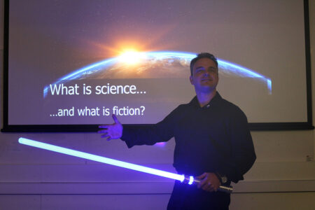Professor Carsten Welsch with a lightsaber in front of a screen with the question "what is science and what is fiction?"