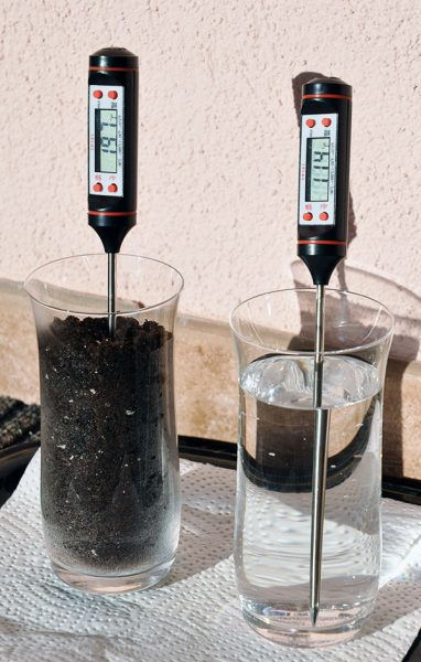Two digital thermometers are held in two containers, one with water and the other with soil. The thermometer in water measures a lower temperature by several degrees.