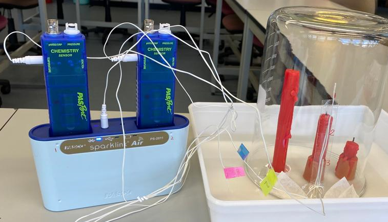 On a table, there is a container which holds the glass dome with the candles, connected to temperature sensors by cables.