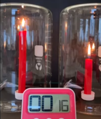 A digital clock sitting on top of a table and two lit candles enclosed in glass jars behind it.